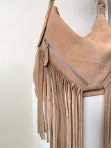 Bolso tipo hobo color tostado, hecho de cuero con flecos largos en el frente y dos bolsillos con cierre. Tiene una manija regulable con hebillas metálicas.