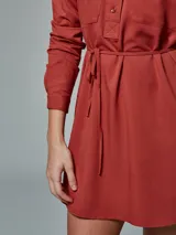 Vestido camisero verde con cuello clásico, mangas largas, cierre frontal con botones, dos bolsillos con solapa en el pecho y cinturón con lazo.