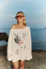 Sweater tejido color crema con cuello amplio que deja un hombro al descubierto. Presenta un estampado con dibujos de estilo boceto en negro y rojo, y detalles de roturas en las mangas.