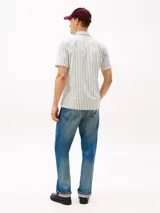 Camisa de manga corta de algodón, color blanco con rayas verticales finas en verde. Presenta cuello clásico con abertura en V y un bolsillo de parche en el pecho con logo bordado.