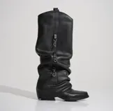 Bota alta de cuero negro con caña arrugada y detalles de cadenas colgantes.