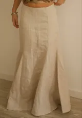Falda larga color beige, de lino, con corte sirena que se ajusta a la cadera y se abre en el ruedo. Presenta costuras verticales que definen la forma y una pequeña abertura lateral en la parte inferior.