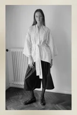 Kimono blanco con rayas verticales negras, confeccionado en tela camisera. Presenta cuello mao, mangas anchas 3/4, bolsillos laterales invisibles y cinturón ajustable desmontable.