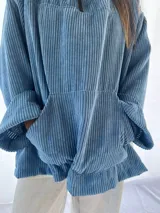 Buzo de pana fina color verde oliva, con diseño de corte holgado y mangas largas. Presenta una textura de cordón vertical característica de la pana y un pequeño parche decorativo en la parte superior de la espalda.