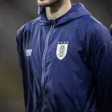 Campera impermeable azul marino con capucha, cierre frontal con cremallera y bolsillos laterales con cierre. Incluye el escudo de la Asociación Uruguaya de Fútbol (AUF) y la bandera de Uruguay en el pecho.