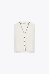 Blusa blanca plisada sin mangas, con cuello redondo y cierre frontal con botones. Presenta vivos negros en cuello, sisas y botonera.