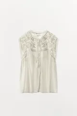 Blusa sin mangas color blanco con cuello alto, bordados y puntillas combinadas a tono, y apliques de abalorios. Cierre frontal con abertura, lazada y botón.