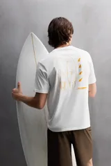 Camiseta blanca de corte holgado confeccionada en tejido de algodón con elasticidad. Tiene cuello redondo, manga corta y estampado en el frente con el logo de la marca y un diseño de tablas de surf.