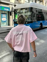 Remera rosa de corte recto con estampado en la espalda que incluye el texto "Itta Tours", "Tour", y nombres de ciudades como Ibiza, Madrid, Andorra, Barcelona y Formentera.