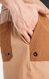Short de baño de calce regular, color camel (marrón claro), con paneles de tela en tono marrón más oscuro en los bolsillos cargo laterales y en la parte superior de los bolsillos delanteros. Posee cintura elástica con cordón negro estampado.
