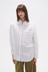 Camisa de algodón blanca de corte holgado, con cuello camisa y manga larga. Presenta un estampado de finas rayas verticales negras y un bolsillo frontal con rayas horizontales.