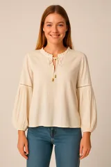 Blusa color crudo con cuello redondo con volados y lazo con detalles dorados, mangas largas abullonadas con detalle de calado.