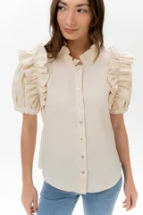 Camisa color beige claro, confeccionada en lino, con cuello alto con volados y mangas cortas abullonadas con volados. Cierre frontal con botones nacarados.