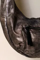 Bolso bandolera de cuero color marrón oscuro con acabado metálico, forro de algodón y herrajes plateados. Tiene correa ajustable y removible, cierre superior y bolsillo frontal con cremallera.