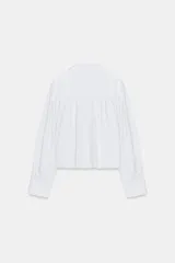 Camisa blanca de popelina con cuello solapa, manga larga abullonada con puño y bajo recto. Cierre frontal con botones ocultos por solapa.