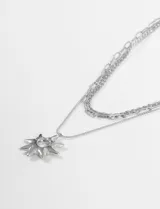 Collar de tres cadenas plateadas de diferente grosor y estilo. La cadena más corta es de eslabones finos, la del medio es de eslabones gruesos y la más larga lleva un dije colgante con forma de sol estilizado y calado.