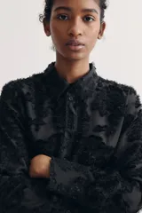 Camisa negra con textura, cuello de pico, botones delanteros, canesú con pliegues en la espalda, mangas largas con hombros caídos, puños abotonados y bajo redondeado.