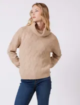 Buzo de punto grueso color beige, con cuello volcado y textura en relieve. Tiene mangas largas de corte hombro caído y puños anchos.