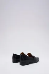 Mocasín negro de cuero liso, con punta cuadrada y hebilla metálica plateada en el empeine. Posee un taco bajo y cuadrado.