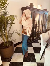 Sweater beige de corderito con capucha y bolsillo frontal de cuero en tonos marrón y ocre. Presenta tajos laterales y cierre parcial con cremallera en el cuello.