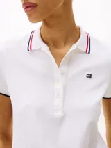 Chomba blanca de corte slim fit, confeccionada en algodón elástico. Presenta cuello y mangas con ribetes en azul y rojo, tapeta de cinco botones y logo de Tommy Hilfiger bordado en el pecho.