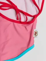 Traje de baño entero rojo con tirantes finos y espalda cruzada. Presenta un parche bordado con un monstruo en la parte inferior. Tiene ribetes en color borgoña y celeste en la parte superior, y ribetes en color naranja y verde en la parte inferior.