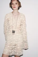 Vestido corto color crudo confeccionado en tejido tipo crochet con diseño geométrico floral. Tiene cuello solapa, abertura con botón en la parte superior y mangas largas acampanadas.