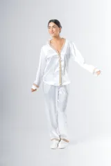 Pijama de satén blanco con detalles de puntilla beige en el escote, puños y ruedo del pantalón. Camisa de manga larga con botones y pantalón recto con bolsillos y elástico en la cintura.