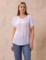 Blusa blanca de gasa con cuello redondo y manga corta japonesa. Presenta apliques de crochet en el cuello, ruedo de las mangas y en el frente.