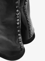 Bota alta de cuero negro con diseño de caña ancha y efecto de solapa que cubre el talón. Presenta detalles de tachas metálicas en la costura posterior, punta redondeada y suela con taco bajo.