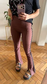 Pantalón de gabardina elastizada color malva o rosado oscuro, de tiro alto y corte acampanado. Presenta un ribete decorativo negro con borde festoneado a lo largo de las costuras laterales exteriores de las piernas.