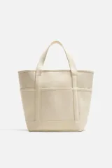 Bolso tipo shopper color crema, fabricado en tejido de algodón. Tiene doble asa corta, compartimento principal con cierre de cremallera, bolsillo interior con cremallera y múltiples bolsillos exteriores.