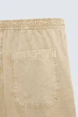 Pantalón de algodón color beige con efecto lavado, corte relajado y pierna ancha. Presenta cintura elástica con ajuste de cordón y cierre frontal con botón, además de bolsillos delanteros y bolsillos traseros de plastrón.
