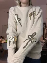 Suéter de punto color beige claro, con cuello redondo y puños y ruedo acanalados. Presenta apliques de moños hechos con lentejuelas doradas y plateadas en el frente y mangas.