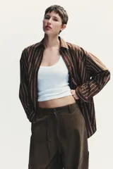 Camisa oversize marrón con rayas verticales blancas, cuello solapa, manga larga con puños y hombros caídos. Tiene un bolsillo de plastrón en el delantero, pinzas en la espalda y cierre frontal con botones.