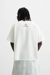 Camiseta blanca de corte holgado con mangas cortas negras y cuello redondo. Presenta estampados frontales con logos y textos en azul y rojo.