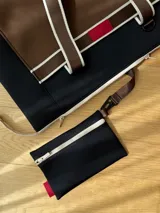 Bolso tipo tote de neopreno color negro y marrón, con cierre superior, bolsillo frontal marrón con logo rojo, bolsillo interno y externo con cierre, doble asa de mano marrón con detalles blancos y opción de correa larga.