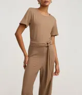 Mono de mujer color marrón topo, con escote redondo, mangas cortas y pantalón ancho. Incluye un cinturón ancho del mismo tejido que se anuda con una hebilla metálica redonda.