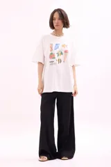 Remera blanca de algodón, corte oversize, con cuello redondo y manga corta. Presenta un estampado frontal con varias reposeras y hamacas de playa, y el texto "LA SIESTA".