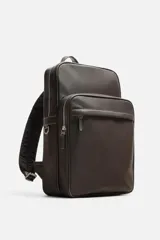 Mochila marrón de cuero sintético con textura grabada. Tiene un compartimento principal con cierre de cremallera, un bolsillo interno para dispositivos electrónicos de hasta 13 pulgadas y un bolsillo interno con cremallera. En la parte delantera tiene un bolsillo con cremallera. La parte posterior es acolchada y tiene una banda de sujeción para equipaje. Tiene un asa de mano y dos asas de hombro ajustables y acolchadas.