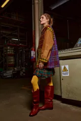 Chaqueta con cuerpo de lana a cuadros en tonos marrón, verde y amarillo. Mangas y cuello de cuero agamuzado color camel. Cierre frontal con cremallera. Falda corta a cuadros escoceses en tonos verde, rojo y azul. Medias amarillas y botas de cuero marrón. 