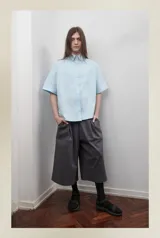 Bermuda gris unisex de corte amplio, con pretina elástica, tiro bajo, bolsillos laterales y cierre lateral. Presenta un pliegue delantero de gran tamaño.