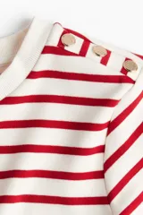 Remera entallada de punto a rayas rojas y blancas con cuello redondo y mangas cortas con botones dorados en los hombros.