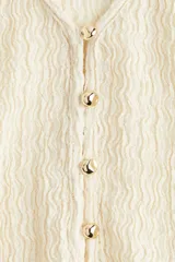 Top color crema de manga larga en punto texturizado. Modelo con escote en V profundo y botones de metal adelante que terminan en abertura en el bajo. Ribete sobrehilado en puños y bajo.