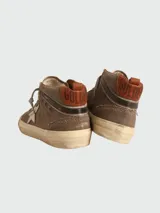 Zapatillas de media caña marca Golden Goose, modelo Mid-Star. Confeccionadas en lona color marrón con efecto desgastado, presentan una estrella de piel blanca en el lateral y detalles de brogue en la puntera y el talón. Cuentan con cordones grises y suela de goma con acabado envejecido.