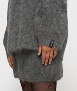 Sweater de punto oversize color gris, confeccionado en una mezcla de lana de alpaca con textura suave y peluda. Presenta cuello en V, mangas largas con hombros caídos y ribetes de canalé. Incluye una etiqueta decorativa en el puño de la manga izquierda.