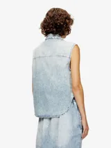 Camisa sin mangas de denim con lavado ácido, cuello camisero y cierre frontal de botones oscuros. Presenta un pequeño pin o aplique en la solapa izquierda.