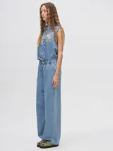 Mono largo de jean celeste con corte holgado, cintura elastizada con cordón ajustable, sin mangas, cuello camisero y cierre frontal con botones. Presenta bordados de motivos playeros en el pecho.