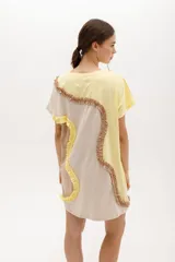 Vestido corto de algodón, con bloques de color en tonos pastel (amarillo claro, beige y rosado pálido) unidos por volados fruncidos en tonos marrón claro y amarillo. Presenta cuello redondo y mangas cortas.