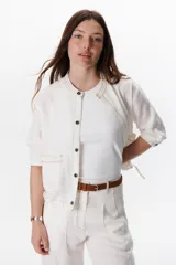 Chaqueta color crudo de lino con cuello redondo, manga tres cuartos, cierre frontal con botones y bolsillos de parche. Detalle de costuras en contraste color marrón.